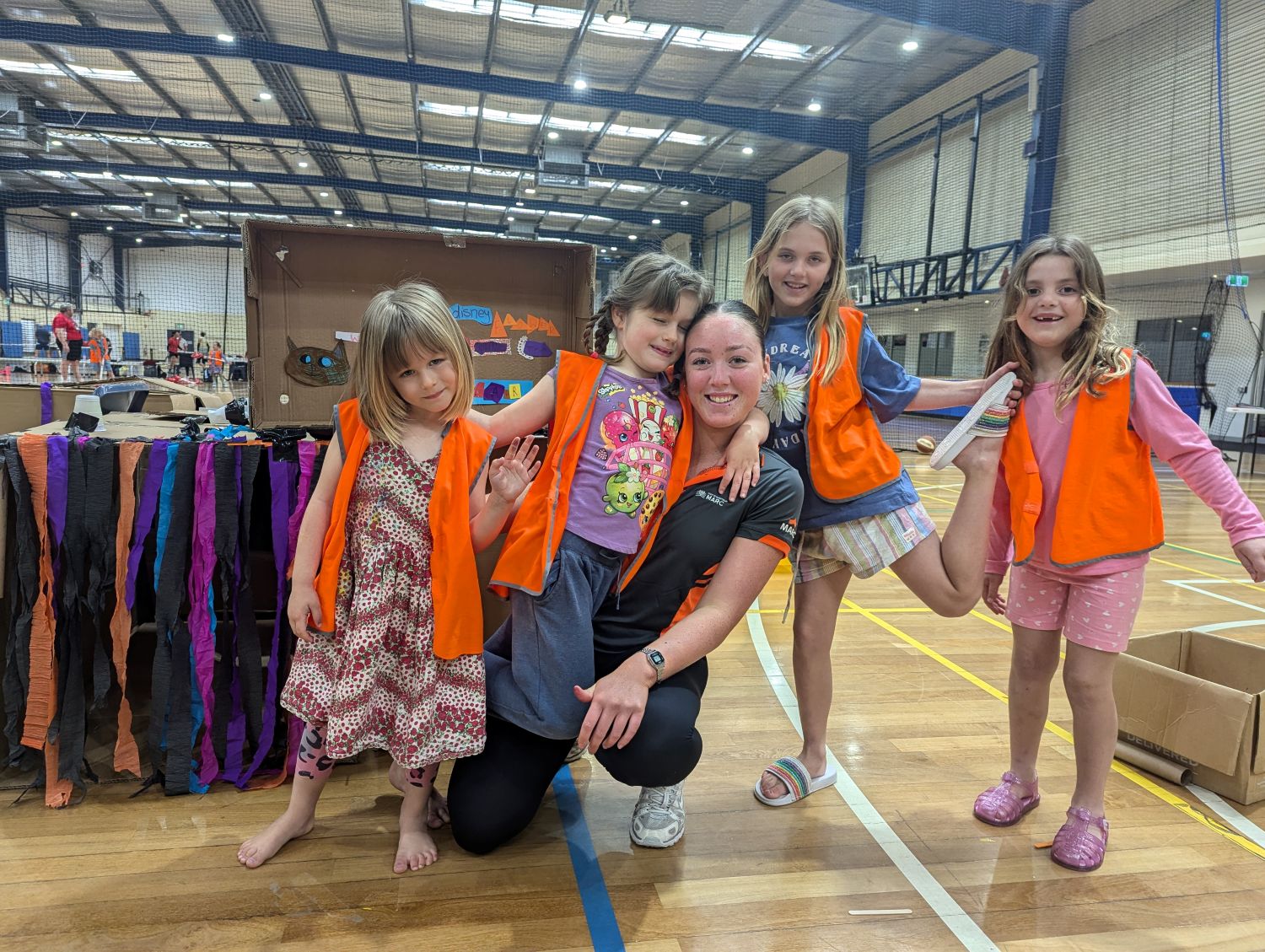 A smiling supervisor poses on an indoor basketball court surrounded by four young girls wearing bright orange high-visibility vests next to a cardboard fort.